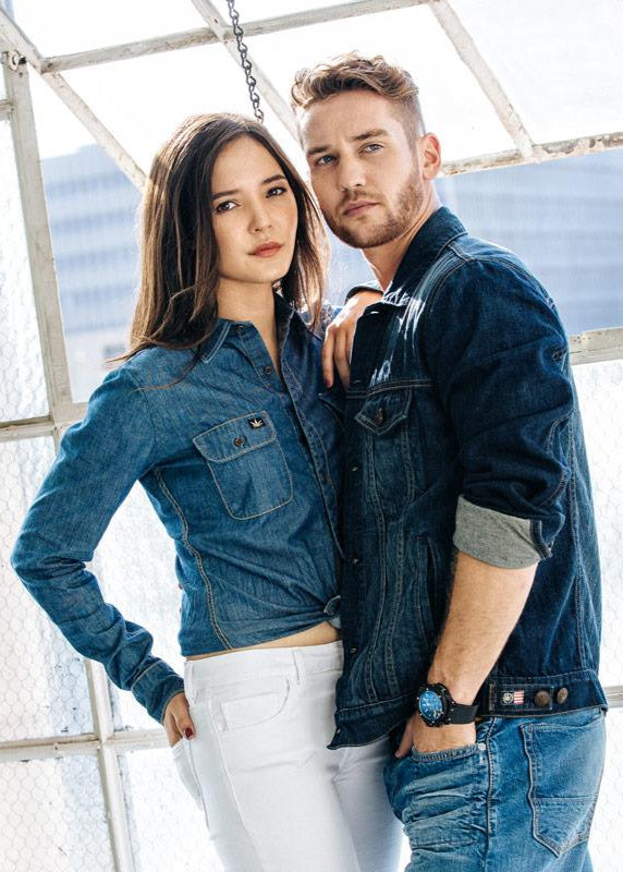 Two people wearing HempBlue sustainable denim clothing standing close together in front of a large window.